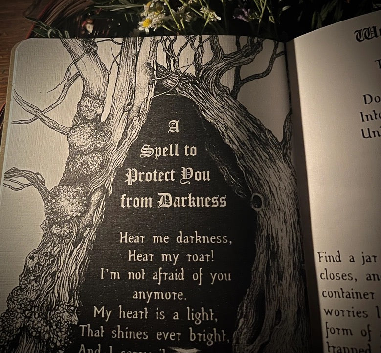 An open book or journal showing a black-and-white illustration of a gnarled tree with the text 'A Spell to Protect You from Darkness' on the left page, photographed indoors with flowers at the top edge.