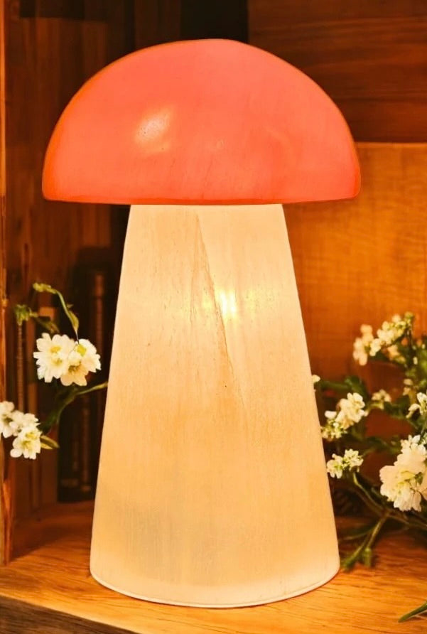 Mushroom-shaped lamp with a red cap and white stem on a wooden surface with flowers in the background.