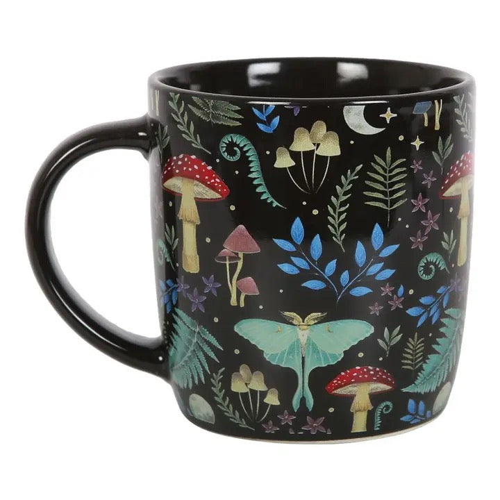 A black ceramic mug decorated with a whimsical woodland pattern featuring green ferns, red and yellow mushrooms, a blue moth, and a crescent moon, shown against a plain white background.