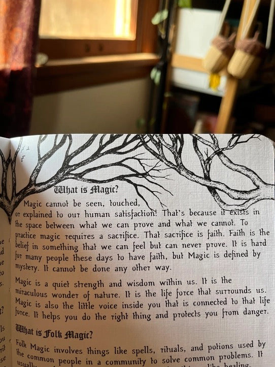 An open book spread with printed text about 'What is Magic?' and 'What is Folks Magic?' and a black tree illustration across the top, photographed in a warm indoor setting with blurred background lights and a person out of focus.