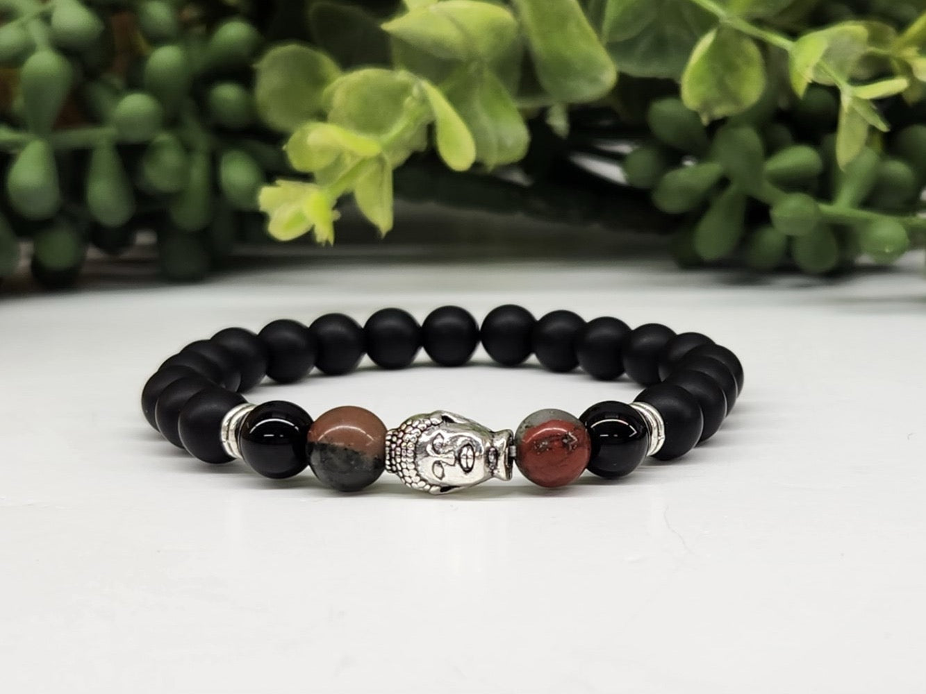 Black beaded bracelet with a silver Buddha figure and red stone beads on a white surface with greenery in the background.