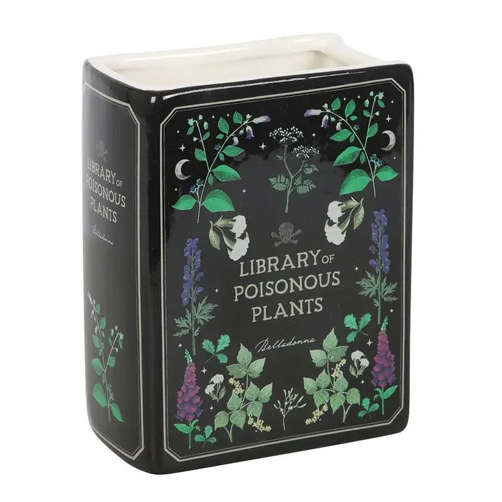 Decorative book-shaped vase with 'Library of Poisonous Plants' design filled with dried flowers on a white surface.