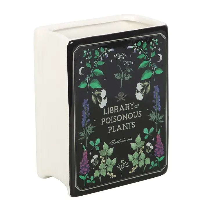 Decorative book-shaped vase with 'Library of Poisonous Plants' design filled with dried flowers on a white surface.