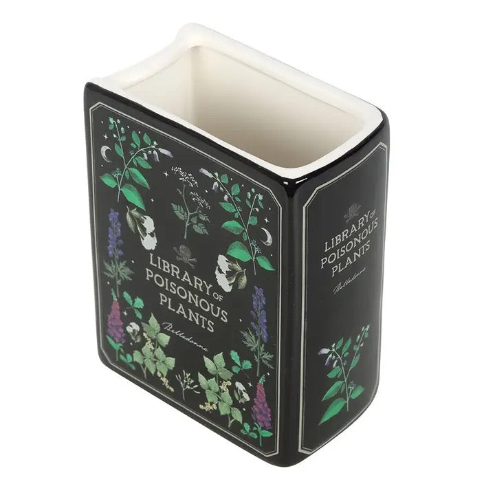 Decorative book-shaped vase with 'Library of Poisonous Plants' design filled with dried flowers on a white surface.