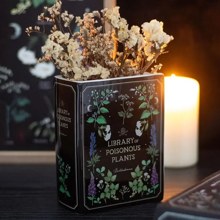 Decorative book-shaped vase with 'Library of Poisonous Plants' design filled with dried flowers on a dark surface.