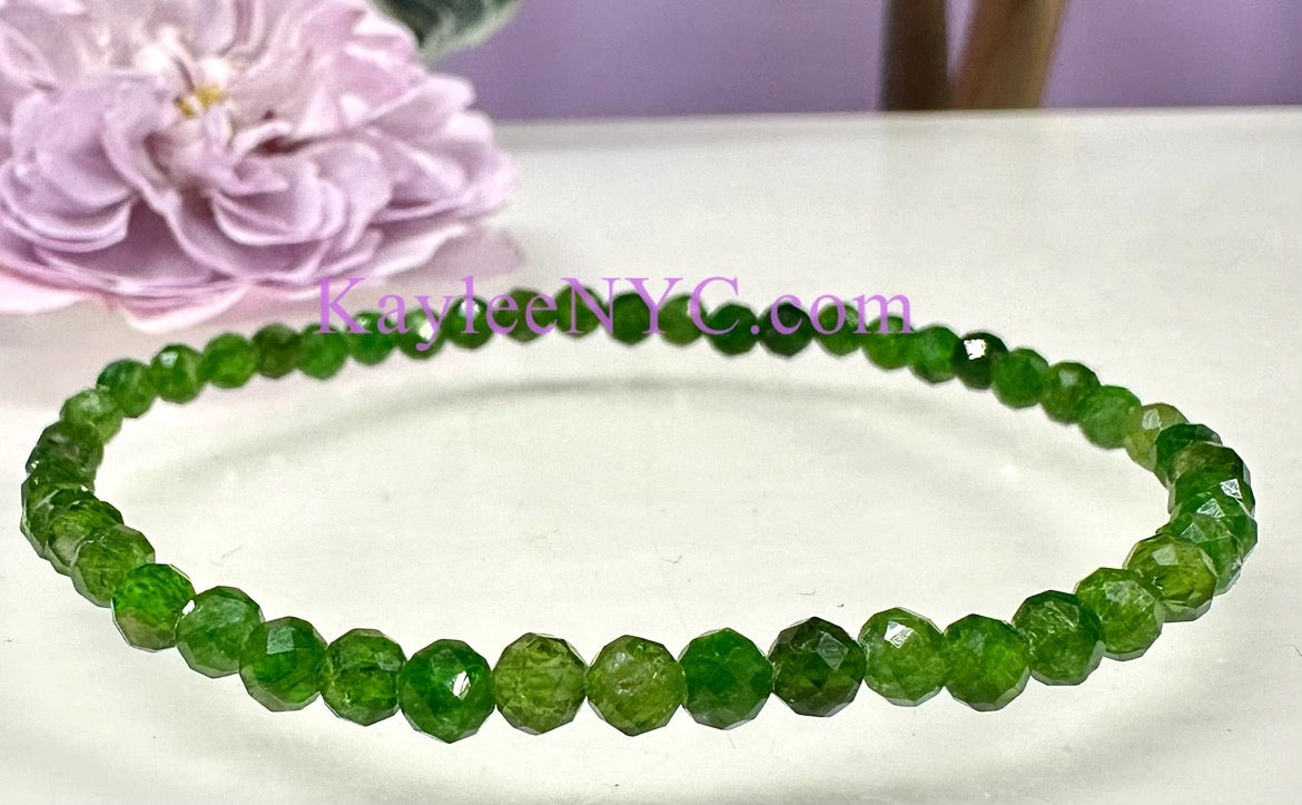 A beaded stretch bracelet made of faceted green beads, displayed on a white surface with a pink flower in the background. A watermark reading 'KayleeNYC.
