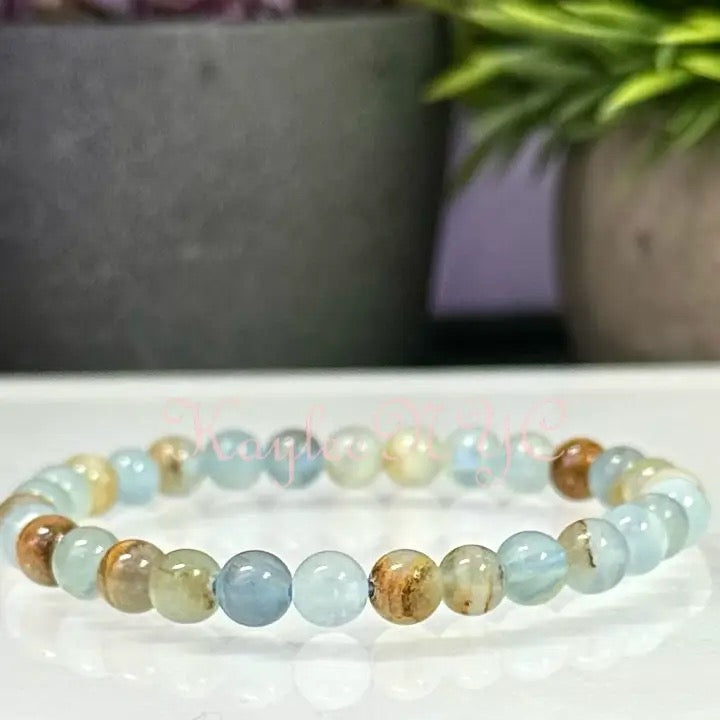 A beaded stretch bracelet made of round light blue, pale green, and brown gemstone beads, displayed on a white reflective surface with a blurred background of potted plants.