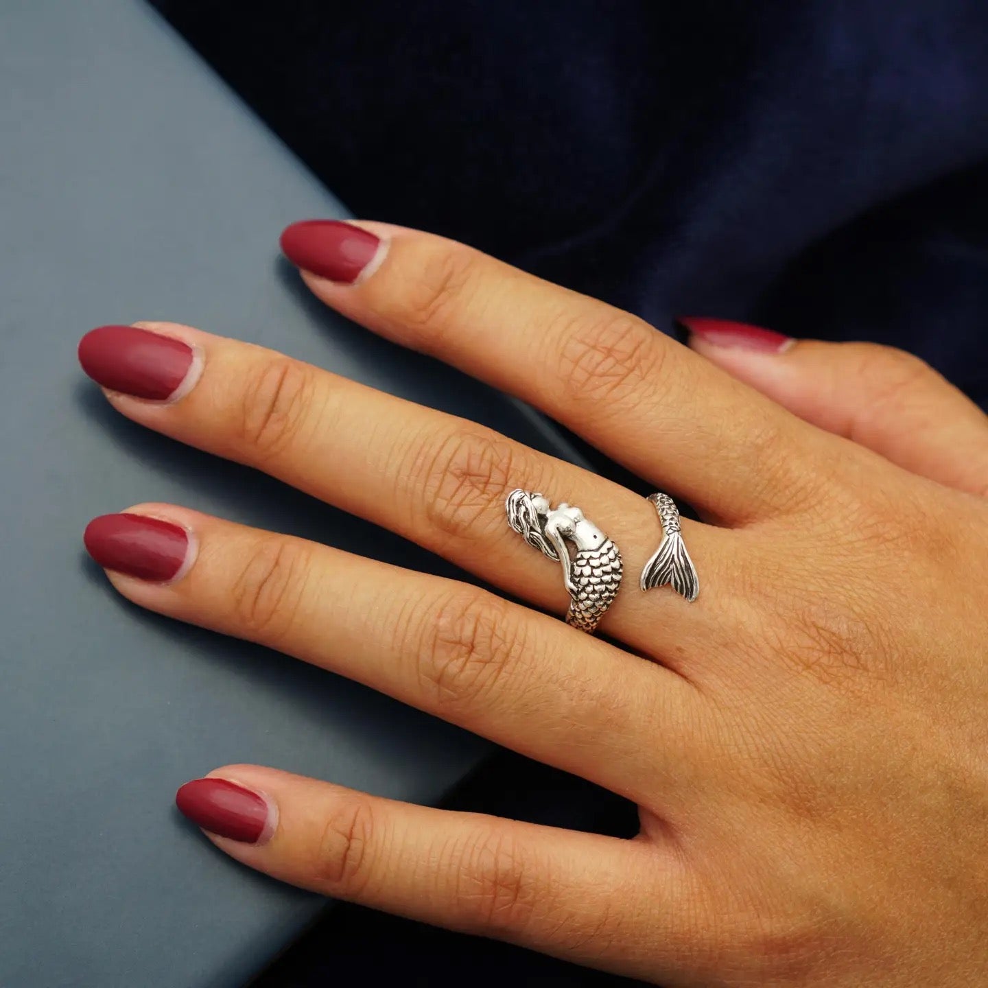 Hand wearing a silver mermaid tail ring with red nail polish on a gray surface