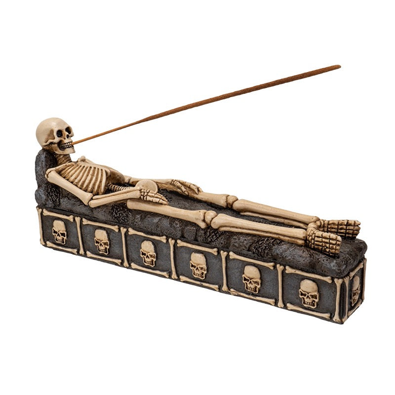Skeleton figure in a coffin with skull designs, holding an incense stick on a white background