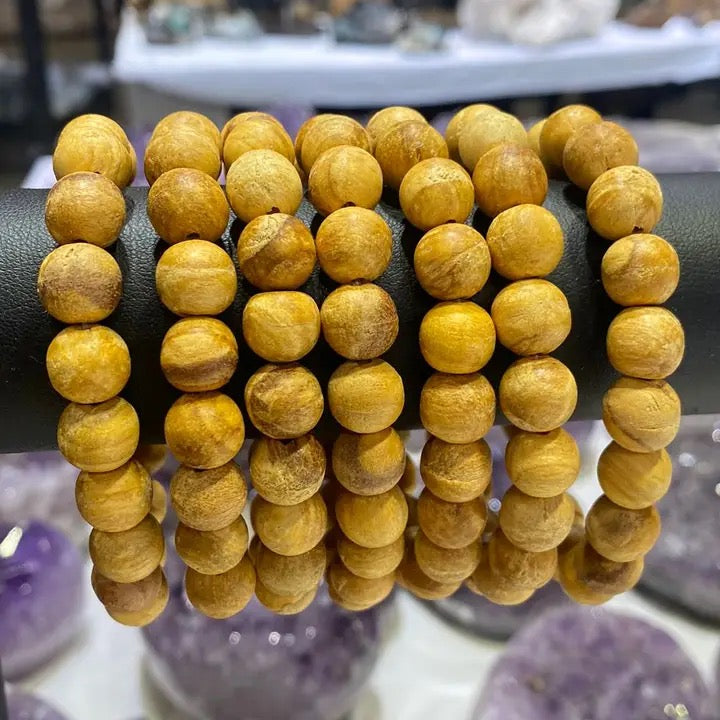 A set of wooden beaded bracelets in a warm golden-brown tone, displayed on a black stand with blurred purple crystal clusters in the background.