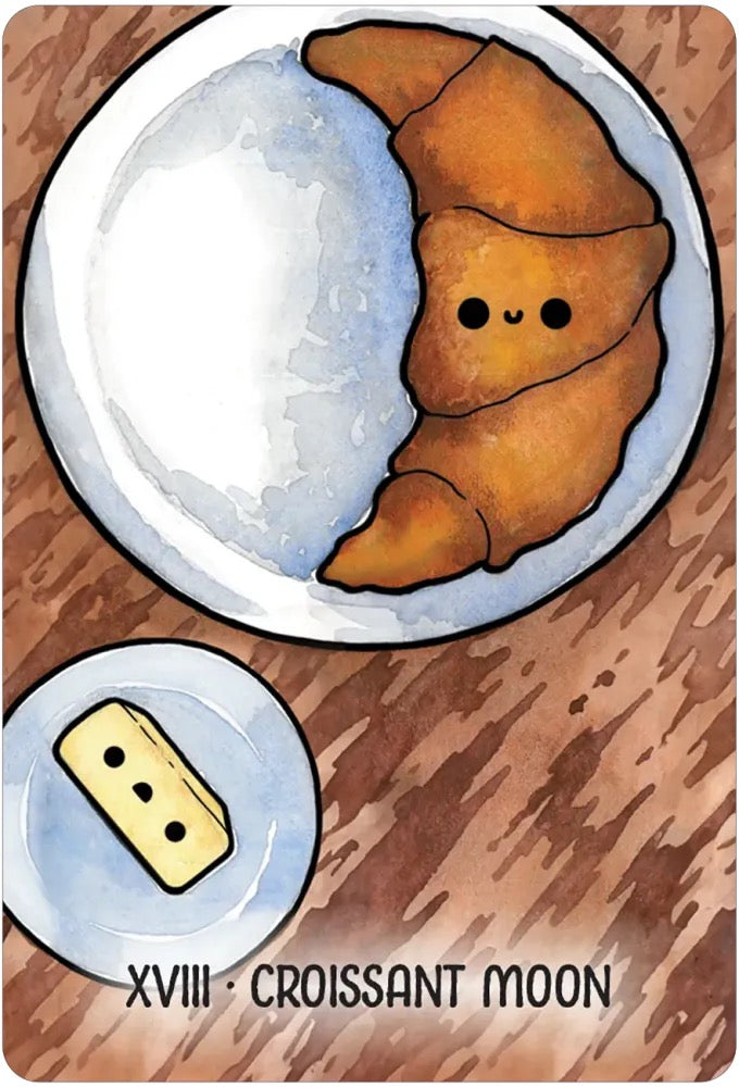 Card featuring a croissant shaped like the moon with a smaller croissant at the bottom, on a wooden surface.