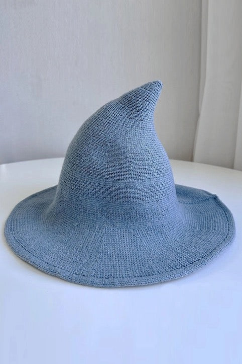 A light blue knit witch hat with a pointed top and wide brim, resting on a white surface against a neutral light gray background.