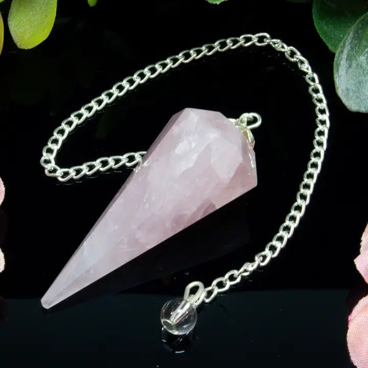 Rose quartz crystal pendulum on a chain against a dark background with green leaves.