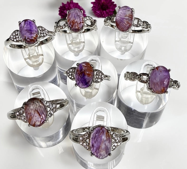 Six silver rings with oval purple amethyst gemstones are displayed on clear acrylic stands against a white background with two purple flowers at the top.