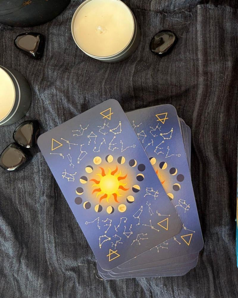 A deck of tarot or oracle cards with a celestial sun and constellation design, styled on a dark textured cloth with candles and black stones.
