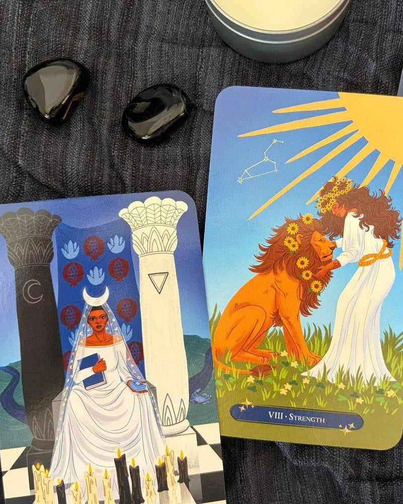 Two illustrated tarot-style cards are laid on a dark textured fabric surface, with two black stones and part of a silver candle tin visible around them. The right card shows a sun, a woman in white, and an orange lion with the text 'VIII - Strength'; the left card shows a woman in white with a crescent moon and blue circular symbols.