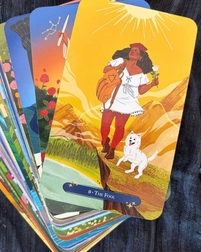 A fanned stack of colorful tarot cards on a dark textured fabric background. The top card is labeled '0 - The Fool' and shows an illustrated figure carrying a backpack with a star emblem, standing outdoors with a white dog under a bright yellow sun.