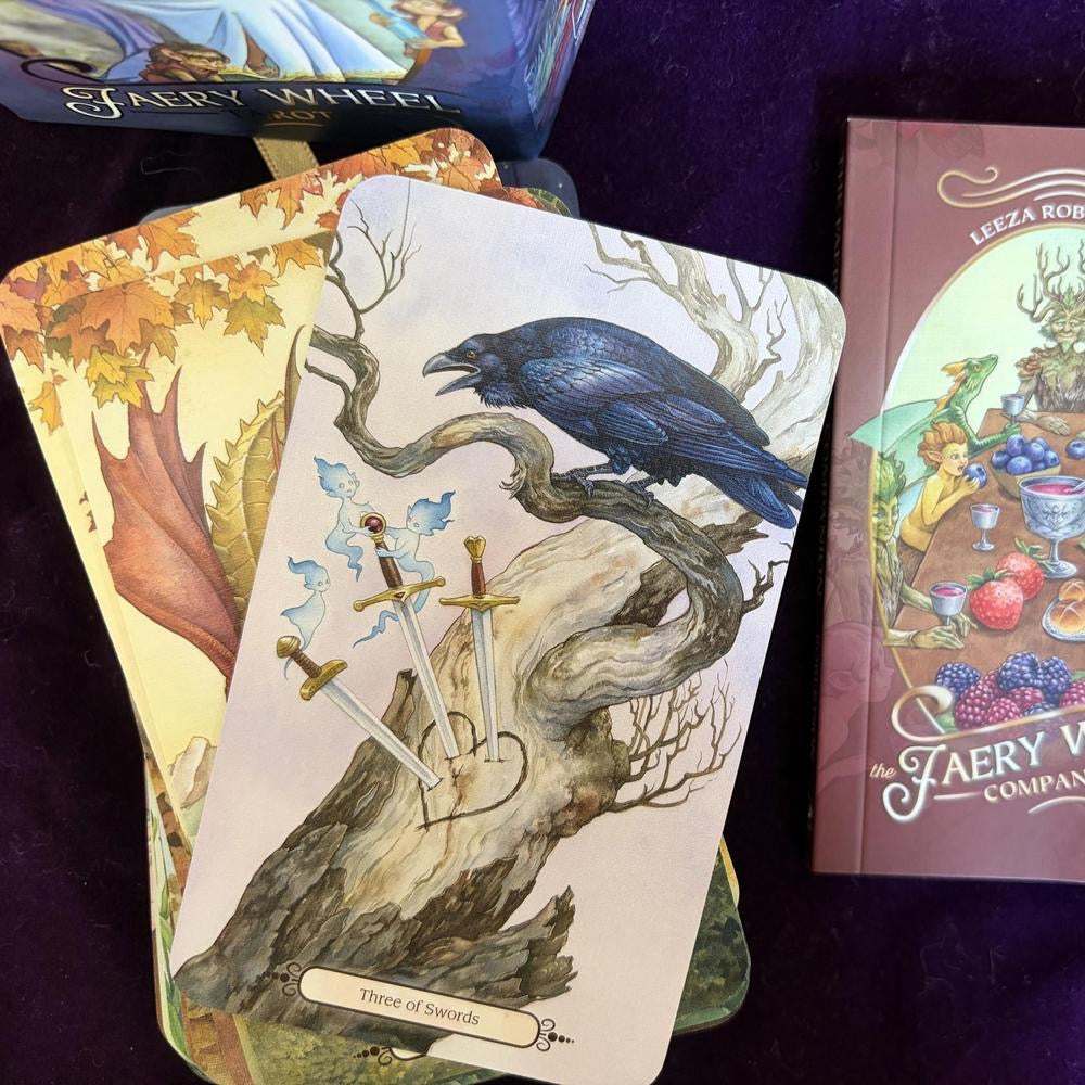 Tarot card with a crow and swords on a purple surface, next to a book titled 'The Faery Wheel'.