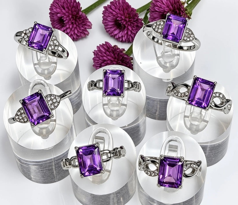 Six silver rings with rectangular purple amethyst gemstones are displayed on clear acrylic stands. The rings feature silver bands decorated with small clear crystals.