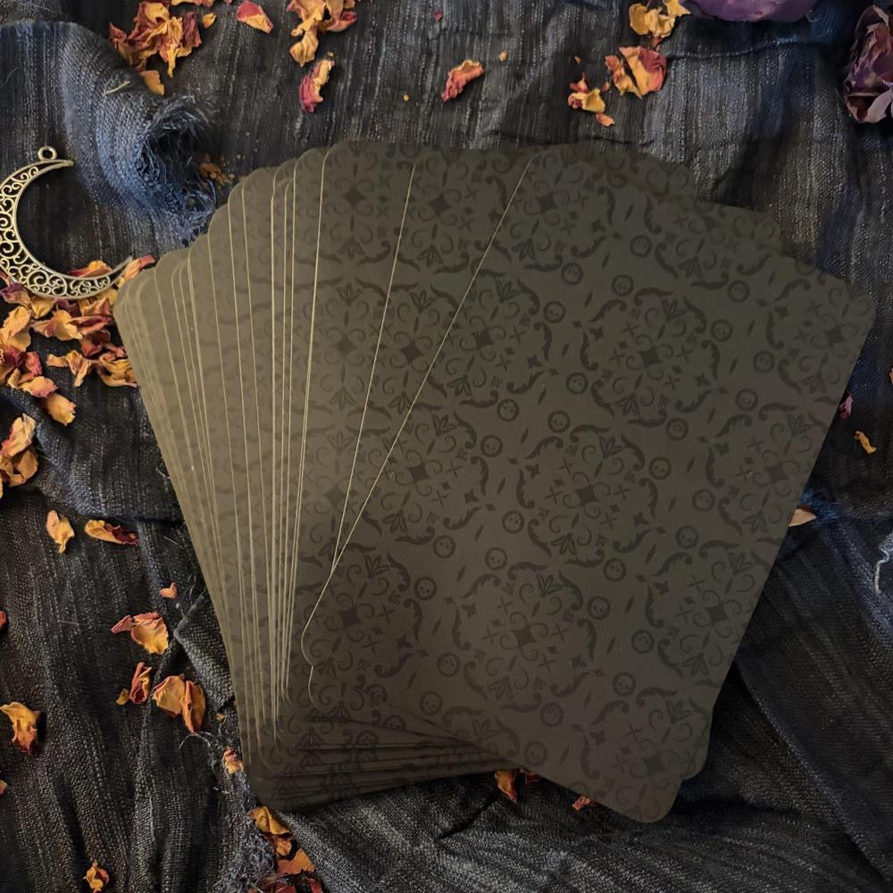 A fan of tarot cards with a dark olive green and black ornate damask pattern, styled on a dark textured fabric with dried orange petals and a silver crescent moon charm.