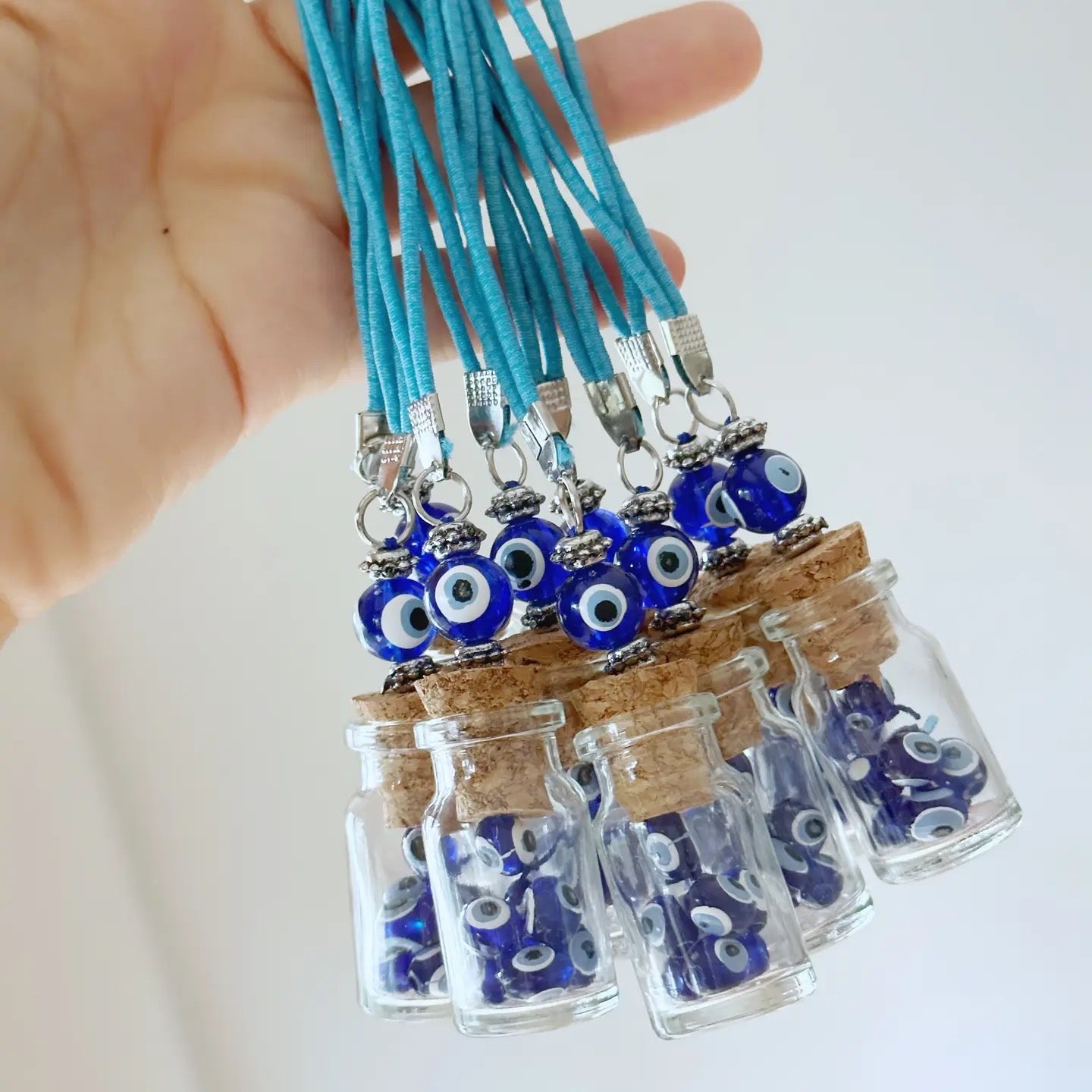 Small glass jars with cork lids containing blue evil eye charms on turquoise strings, held by a hand.
