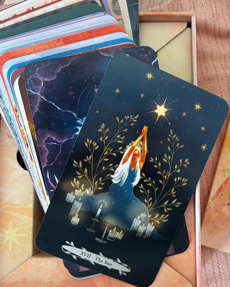 A tarot card deck fanned out in a box, with the top card showing 'XVIII - The Star' and an illustration of a woman reaching toward a glowing star among golden trees on a dark navy background.