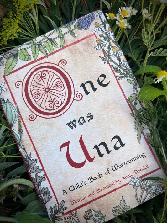 The book 'One was Una' by Annie Dowbell lies on a bed of green garden foliage and small white flowers. The cover features a vintage, aged parchment design with botanical illustrations and elegant typography.