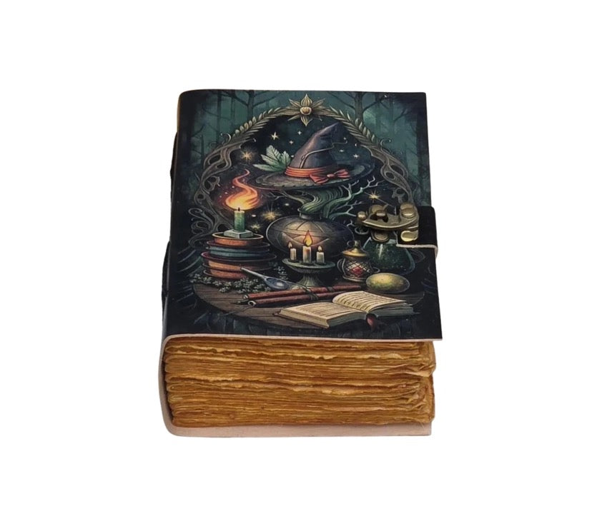 A Leather journal with an illustrated cover depicting a witch's cauldron, potion bottles, a lit candle, an open book, and a pointed hat, set against a dark forest background. The journal has a thick, textured brown faux-leather spine and is shown on a plain white background.