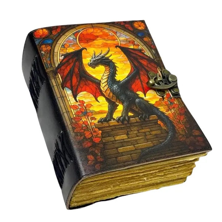 A Leather  journal or notebook featuring a detailed fantasy illustration of a black dragon with red wings perched on a brick wall under a stained-glass-style arch. The cover has a dark textured background, gold-toned page edges, and a small antique-style metal clasp on the right side.
