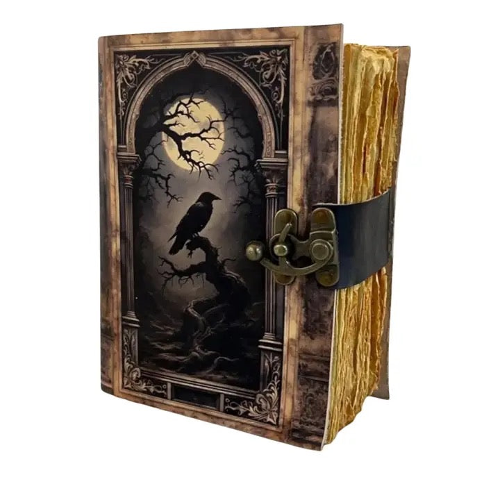 A gothic-style Leather journal with a dark illustration of a raven under a full moon on the cover, featuring a black leather strap and antique brass clasp holding the gold-toned pages.