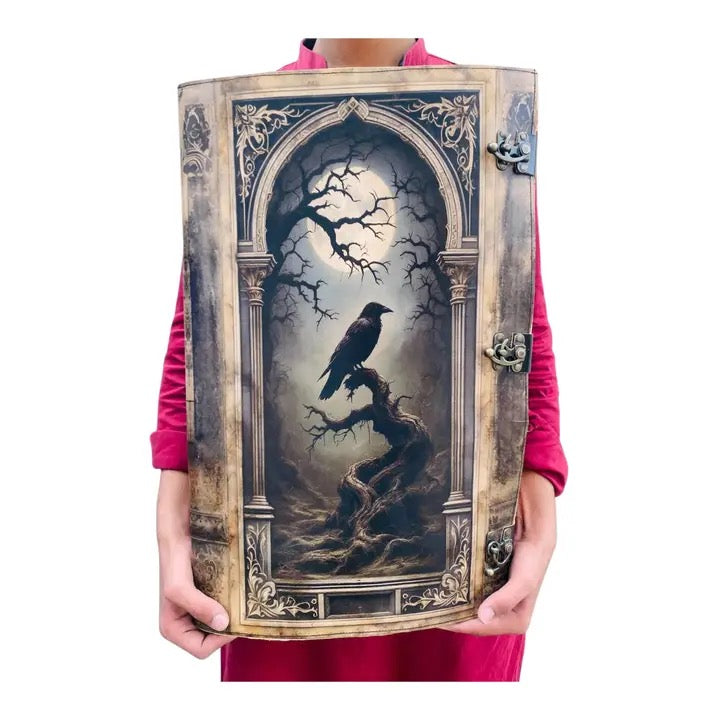 A person wearing a bright pink top holds up a large Leather Journal featuring a gothic illustration of a raven perched on a gnarled tree branch under a pale moonlit sky, framed by ornate columns and arch details.