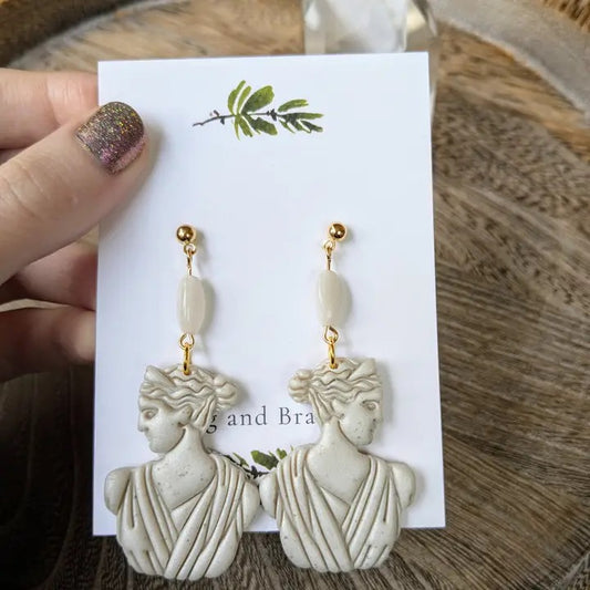 Pair of earrings on a card held by a hand with glittery nail polish on a wooden surface.