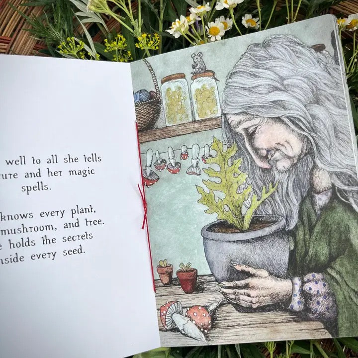 An open illustrated book laid on grass with flowers around it. The left page has partial black text, and the right page shows a detailed drawing of a woman tending potted plants at a table, with jars and small plants in the background.