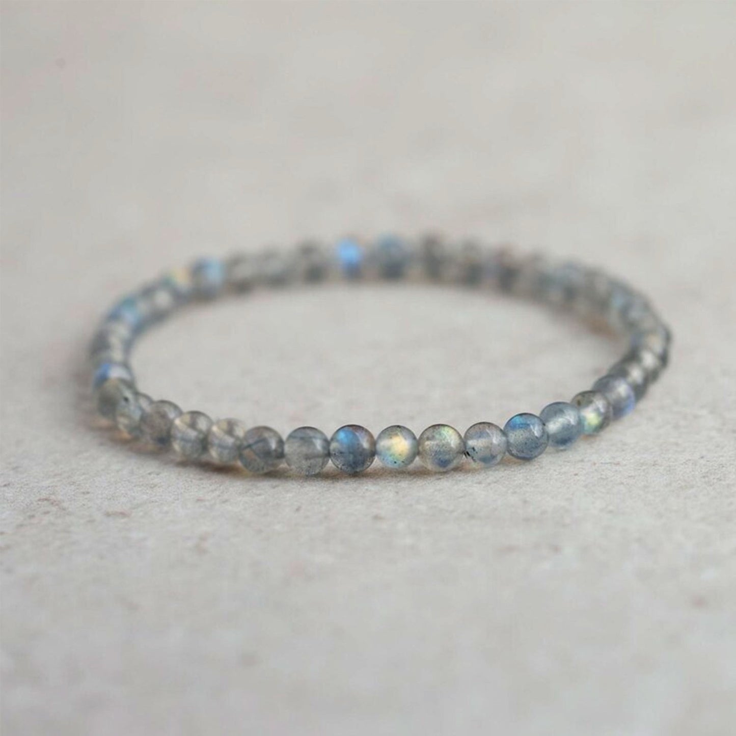 A delicate beaded stretch bracelet made of small, round gray-blue labradorite beads with a subtle iridescent sheen, resting on a light beige textured surface.
