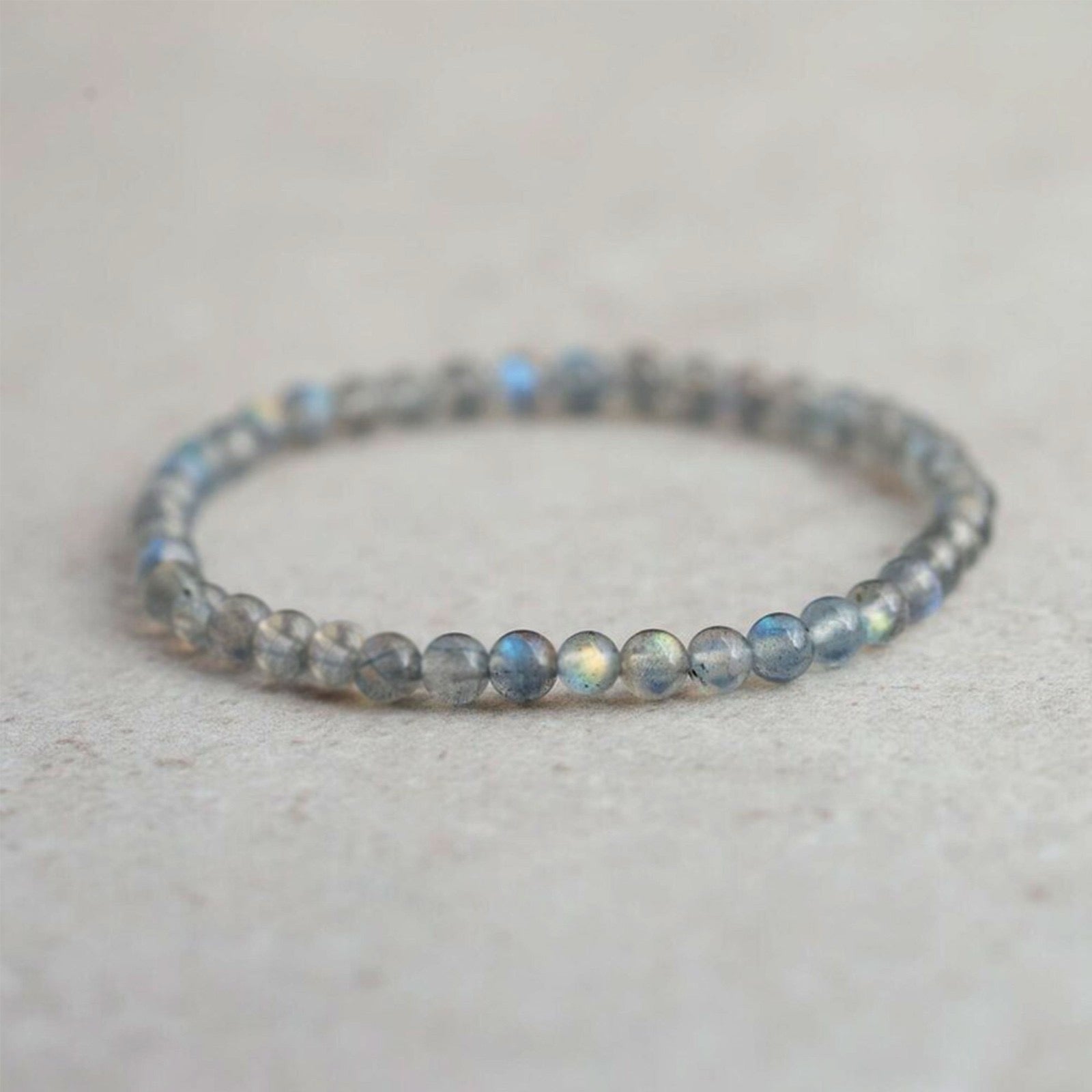 A delicate beaded stretch bracelet made of small, round gray-blue labradorite beads with a subtle iridescent sheen, resting on a light beige textured surface.
