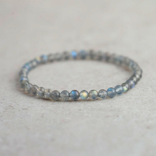 A delicate beaded stretch bracelet made of small, round gray-blue labradorite beads with a subtle iridescent sheen, resting on a light beige textured surface.