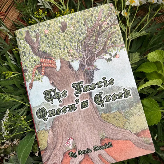 A book titled "The Furry Queen's Creed" by Annie Dowell, featuring an illustrated tree with a red-and-white striped tail and a furry creature on the cover, photographed lying on green garden foliage.