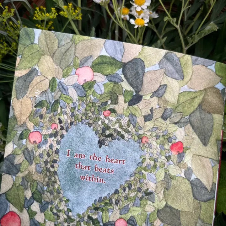 A square printed book page featuring a watercolor-style floral wreath with a pale blue heart in the center and the text 'I am the heart that beats within.' The item is photographed outdoors among green leaves and small white/yellow flowers.