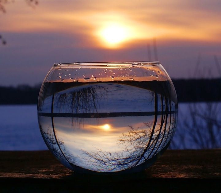 A clear glass fishbowl or decorative bowl filled with water sits outdoors on a wooden surface at sunset, reflecting the sky, water, and tree branches inside the bowl.