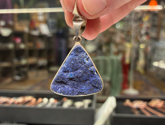 Triangular blue stone pendant held by a hand with a blurred jewelry store background