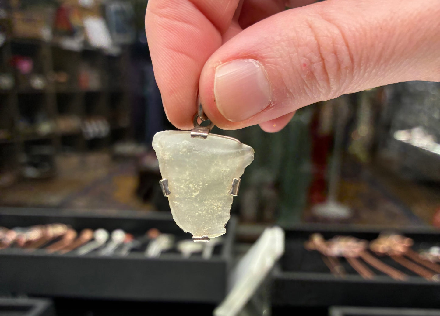Hand holding a clear stone pendant with a blurred background of jewelry items.