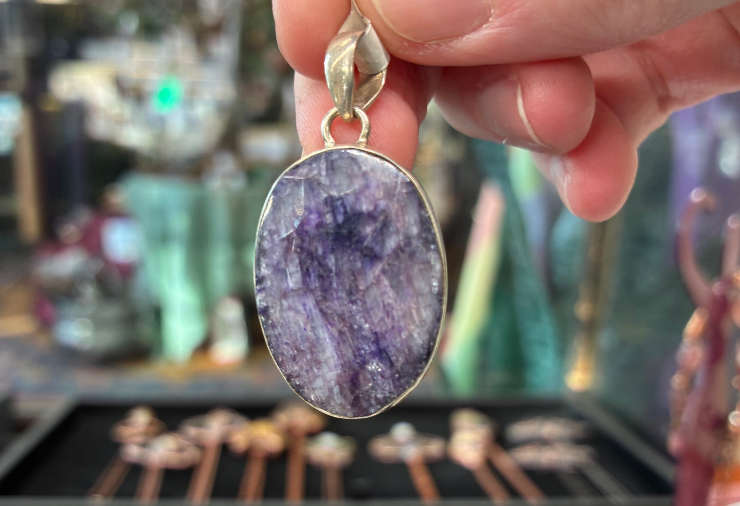 Hand holding a purple amethyst pendant with gold setting against a blurred jewelry store background.