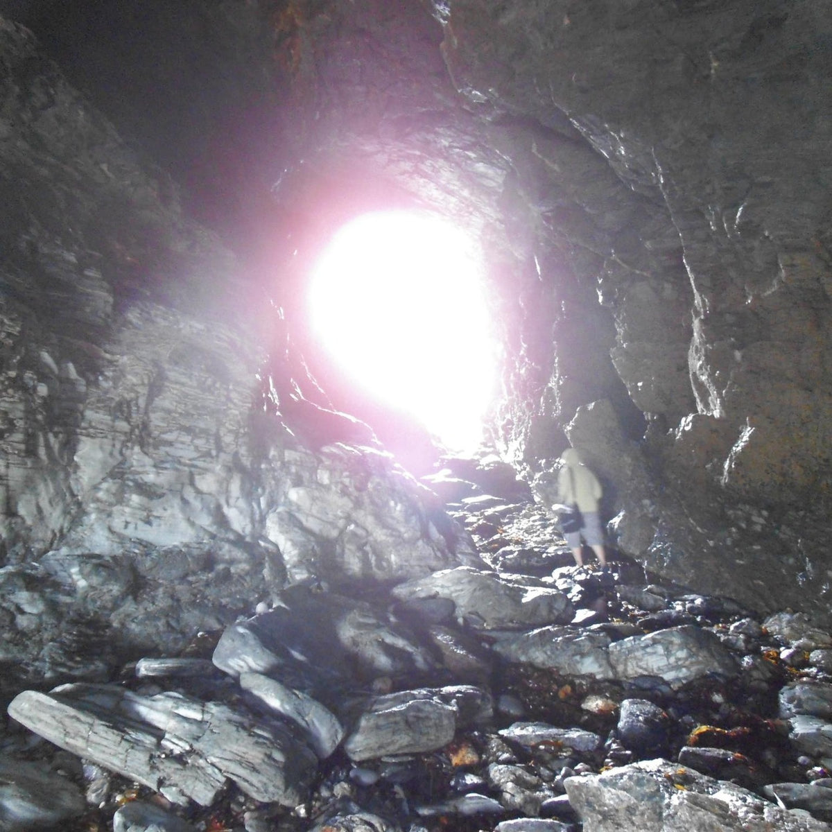 A person standing inside a rocky cave or tunnel with a bright white light glowing from the opening in the distance.