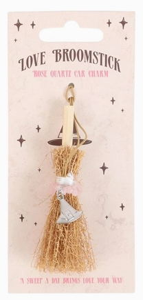 A packaged decorative broom charm on a printed backing card. The charm consists of a small wooden handle, a bundle of tan raffia-like bristles, a miniature silver bell, and a pale pink ribbon.