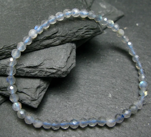 A beaded stretch bracelet made of faceted light blue and white glass beads with subtle iridescent blue flashes, arranged in a circle on dark textured slate stones.