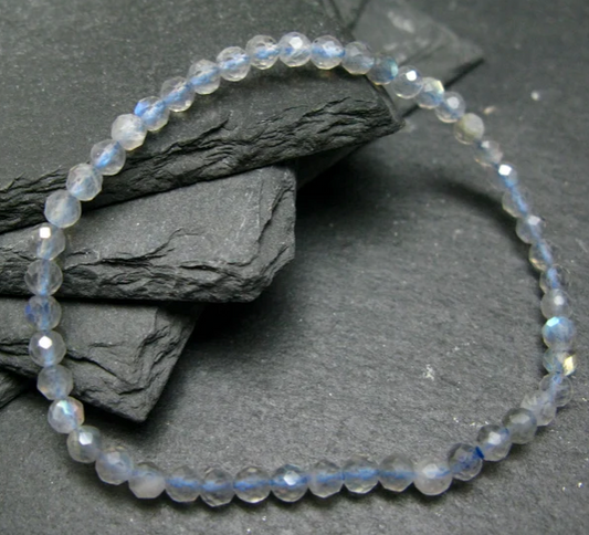 A beaded stretch bracelet made of faceted light blue and white glass beads with subtle iridescent blue flashes, arranged in a circle on dark textured slate stones.