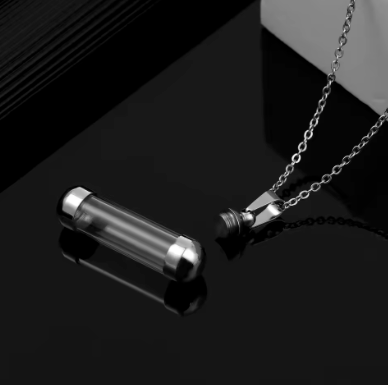 A silver-toned cylindrical pendant necklace with a transparent glass or acrylic tube section, shown on a black reflective surface with its chain extending toward the upper right.