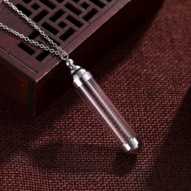 A silver-toned cylindrical pendant necklace with a transparent glass or acrylic vial body, resting on a dark brown textured surface next to a carved wooden block.