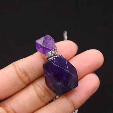 A hand holding a two-tiered purple crystal pendant necklace with a silver-toned metal cap and chain against a dark background.