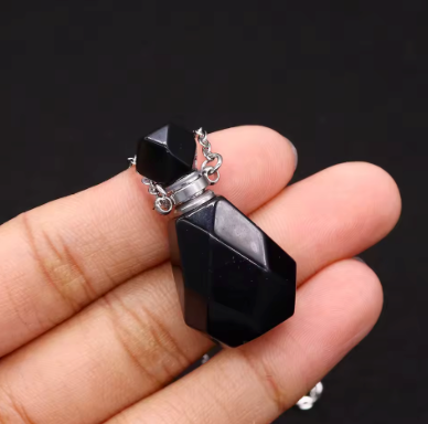 A faceted black glass perfume bottle pendant with a silver-toned chain, held in a person's hand against a dark background.
